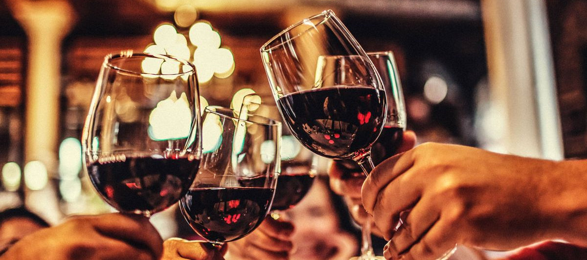 A long, horizontal banner image of a wine collection on a marble countertop, featuring various red, white, and sparkling wine bottles, with glasses of wine alongside them. In the background, there are blurred oak wine barrels and warm lighting.