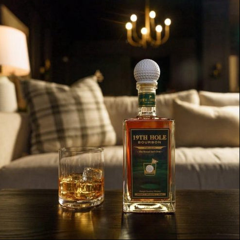 Bottle of 19th Hole Bourbon with a golf ball on top, next to a glass of whiskey on a table.