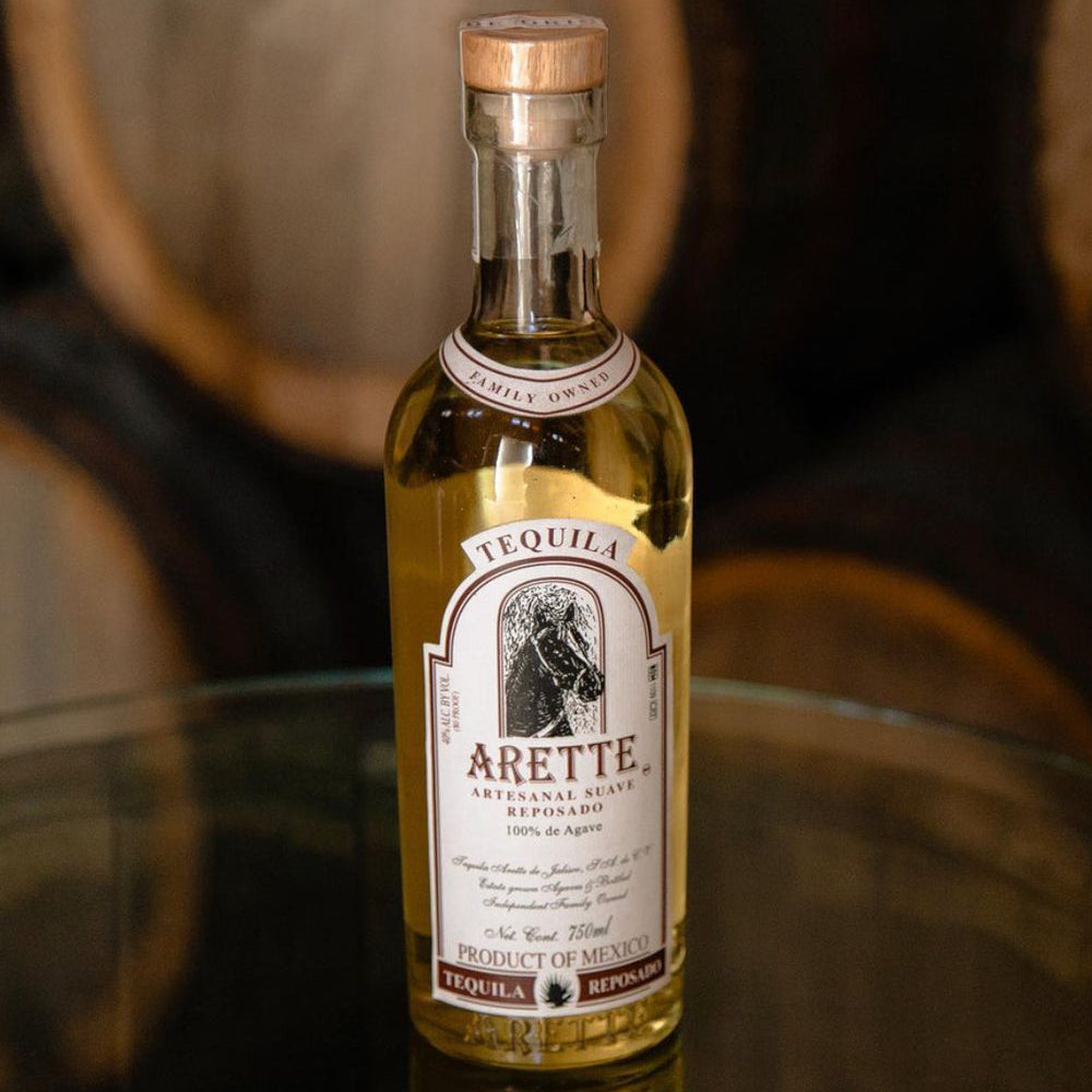 Bottle of Arette Tequila on a reflective surface with wooden barrels in the background