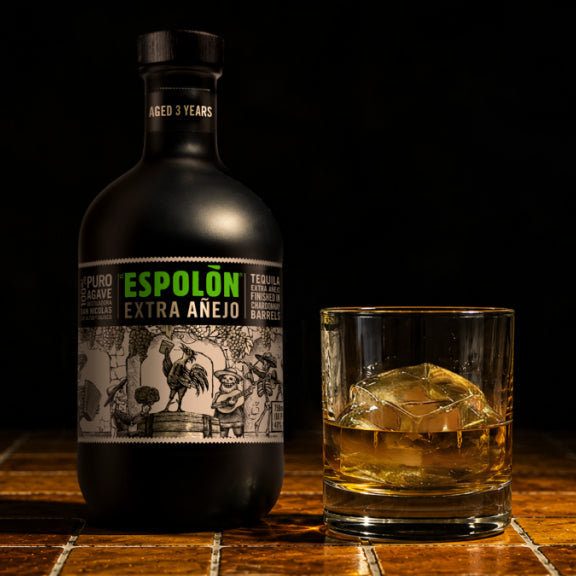 A bottle of Espolón Extra Añejo aged 3 years rum and a glass of amber rum with ice on a dark wooden surface against a black background.