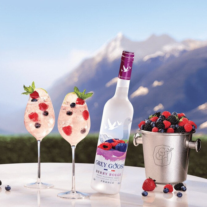 Two glasses of a berry-themed cocktail with a bottle of Grey Goose vodka and a bucket of berries against a mountainous background.
