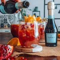Person pouring a drink into a glass with pomegranate seeds and oranges on a wooden table.