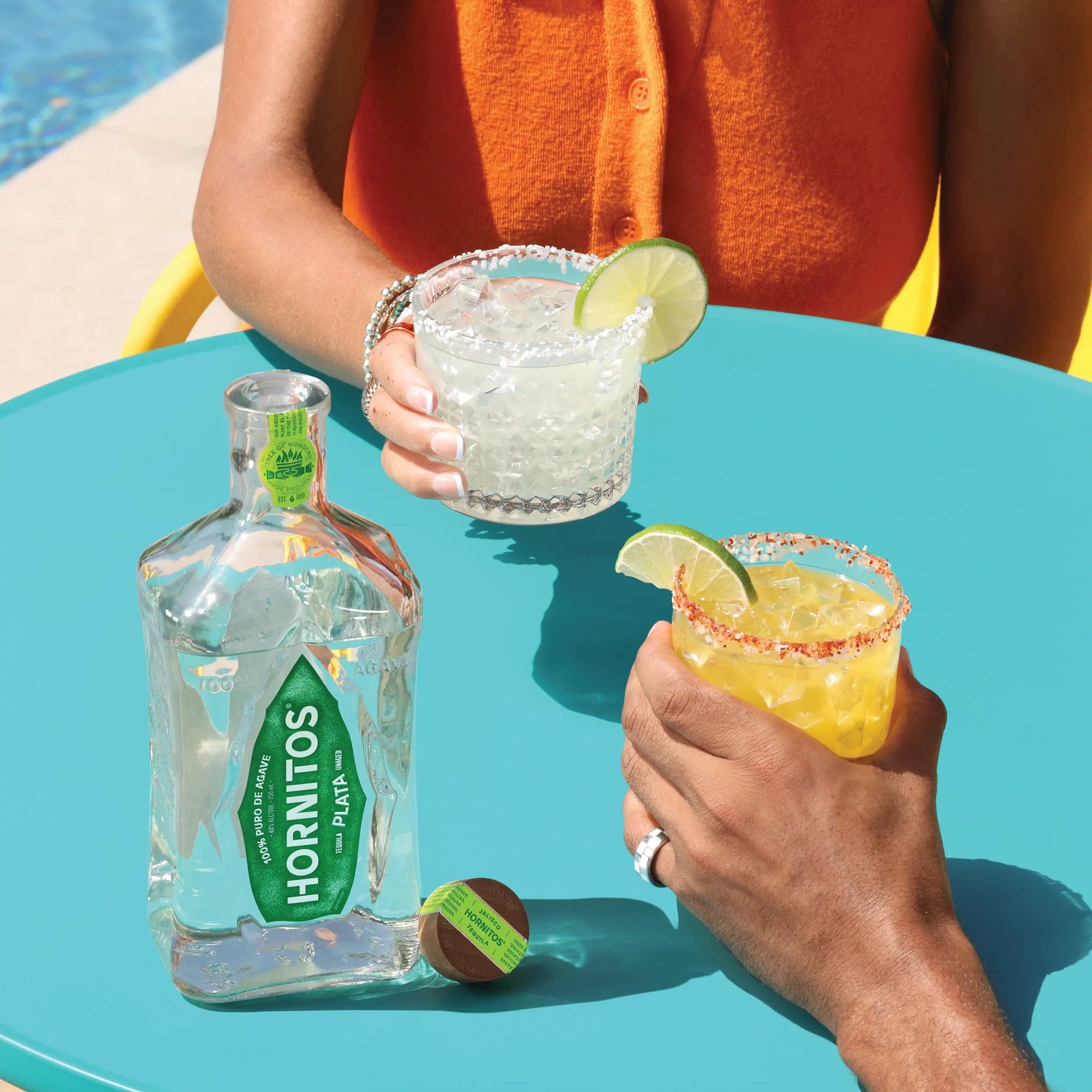 Two people enjoying margaritas with a bottle of Hornitos tequila at a poolside table.