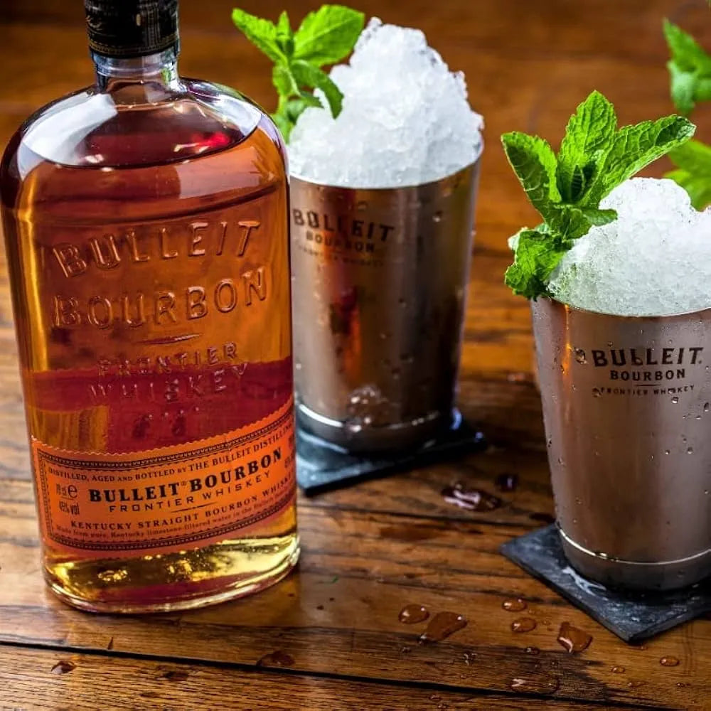 Bulleit Bourbon bottle with two metal cocktail cups filled with crushed ice and mint, served on a wooden bar surface.