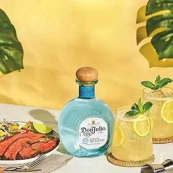 Don Julio Blanco Tequila bottle on a summer table with citrus cocktails and grilled steak, surrounded by tropical leaves.