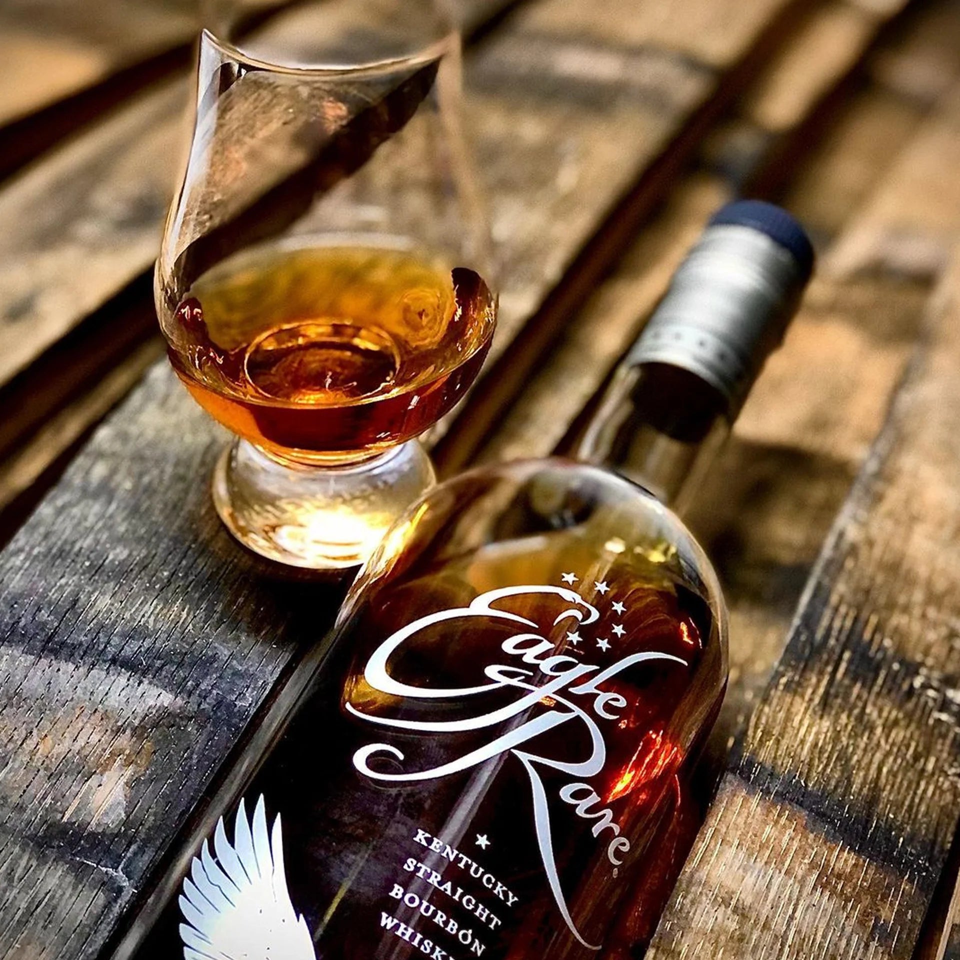 Eagle Rare Kentucky Straight Bourbon Whiskey bottle with a glass pour, displayed on rustic wooden barrel slats, highlighting rich amber color.