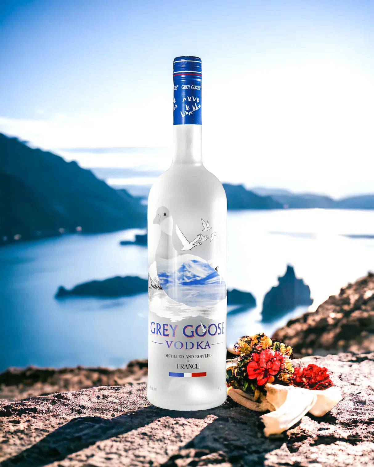 Grey Goose Vodka bottle placed on a scenic cliffside with a mountain lake in the background and red flowers in the foreground, highlighting its French elegance and natural purity.