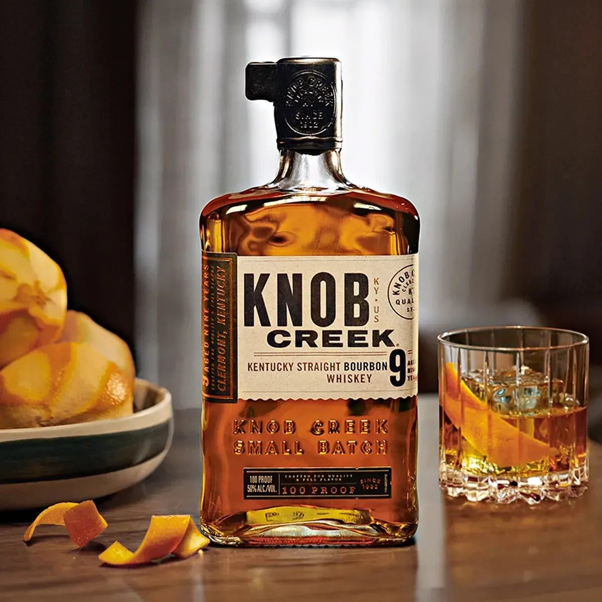 Knob Creek Small Batch 9 Year Bourbon Whiskey bottle on a wooden table with orange peel garnish and cocktail glass.