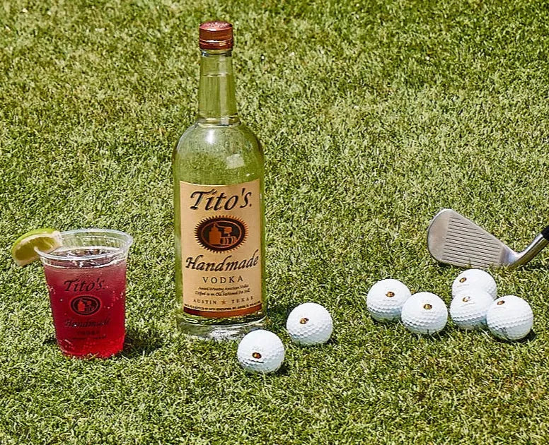 Tito’s Handmade Vodka bottle on a golf course with cocktail, golf balls, and a wedge club — a refreshing outdoor lifestyle scene.