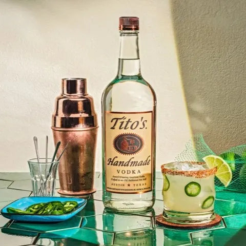 Tito’s Handmade Vodka bottle with a spicy jalapeño and lime cocktail, copper shaker, and fresh garnishes on a vibrant tiled surface.