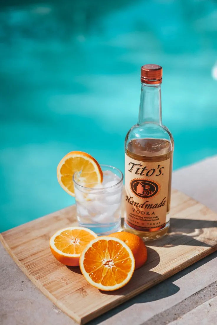Tito’s Handmade Vodka bottle with a citrus cocktail and sliced oranges by a poolside on a wooden board — bright and summery lifestyle scene.