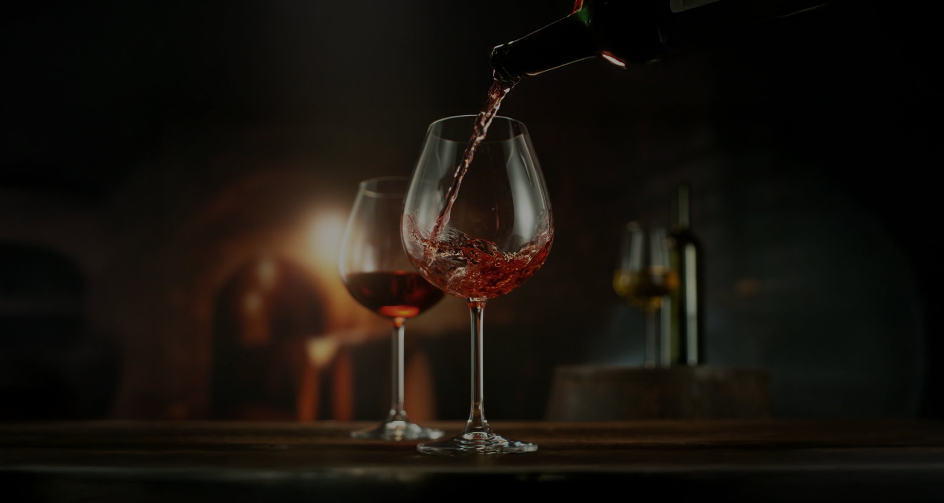 A long, horizontal banner image of a wine collection on a marble countertop, featuring various red, white, and sparkling wine bottles, with glasses of wine alongside them. In the background, there are blurred oak wine barrels and warm lighting.