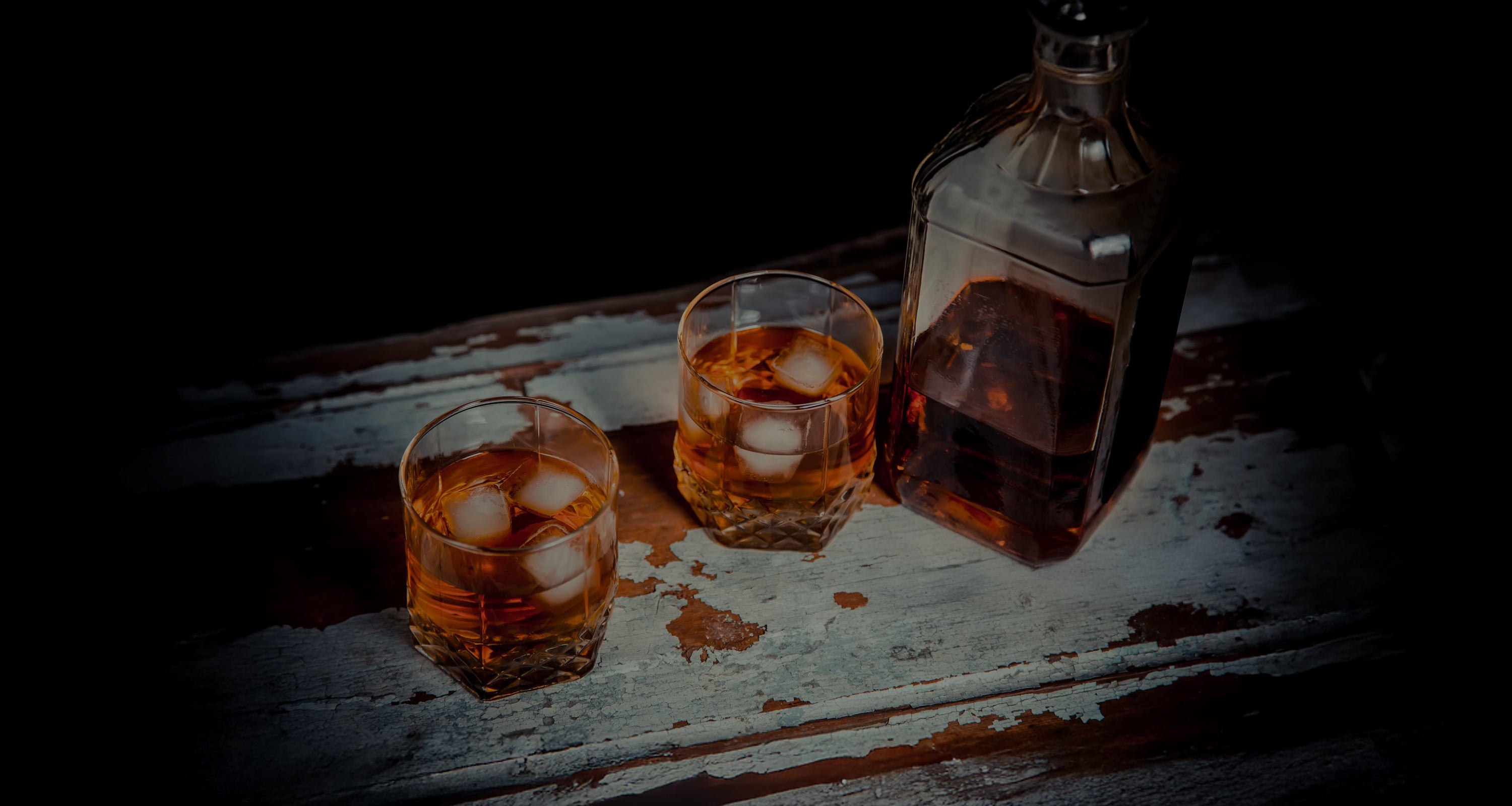 A professional photograph of various whiskey bottles on a wooden table in a whiskey cellar, with barrels in the background.