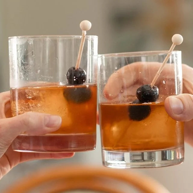 Two hands holding glasses of an Old Fashioned cocktail with cherries on toothpicks against a blurred background.