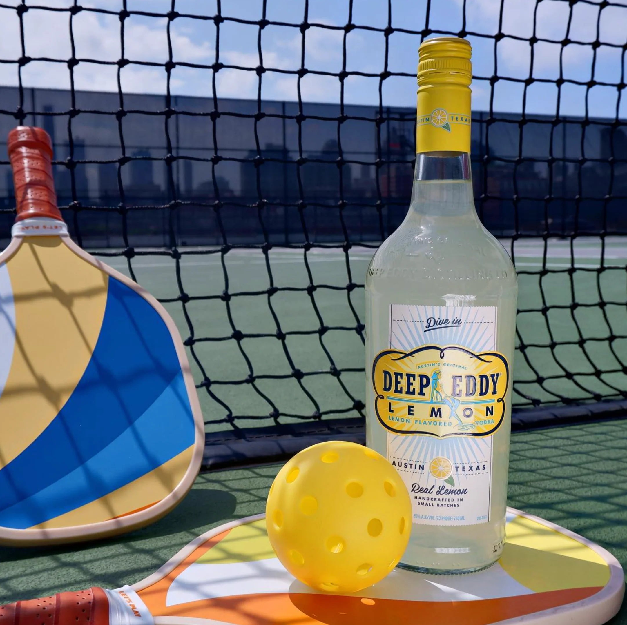 Bottle of Deep Eddy Lemon vodka on a paddle with a pickleball on a court.