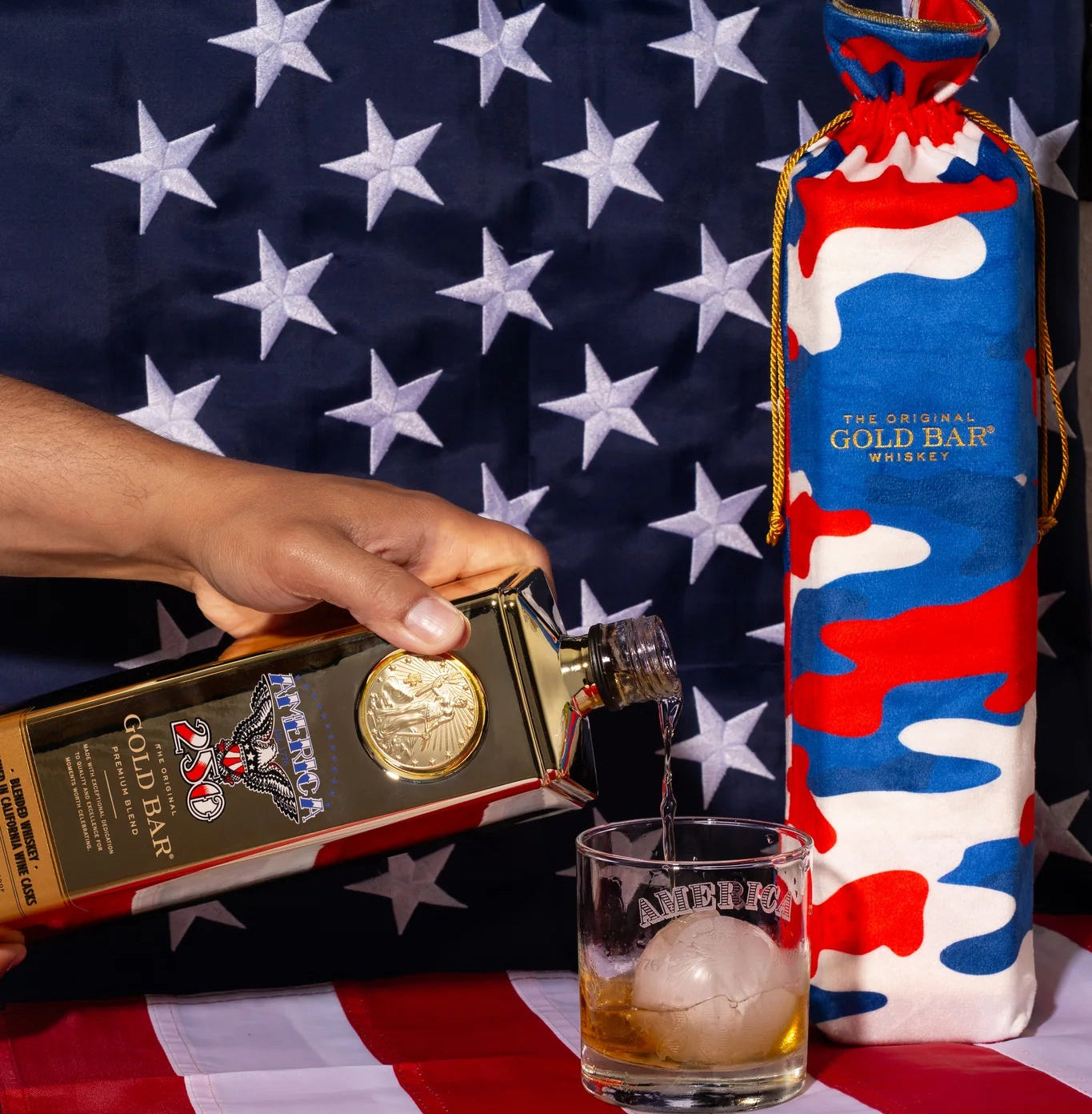 Person pouring whiskey into a glass with a Gold Bar Whiskey bottle and a wrapped gift on an American flag-themed tablecloth.