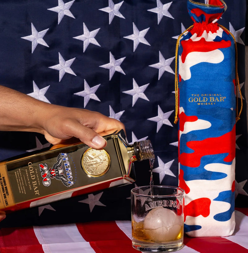 Person pouring whiskey into a glass with a Gold Bar Whiskey bottle and a wrapped gift on an American flag-themed tablecloth.