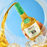 Henny Rita bottle with green cap and label held over a glass of yellow liquid with ice cubes.