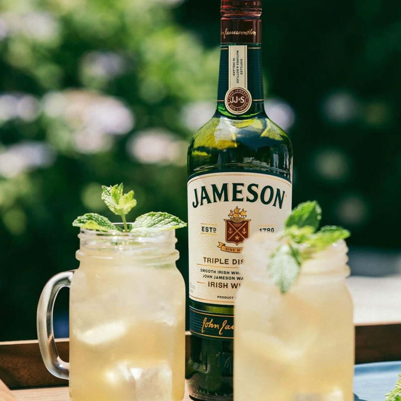 Jameson Irish Whiskey bottle with two glasses of lemonade on a wooden surface outdoors.