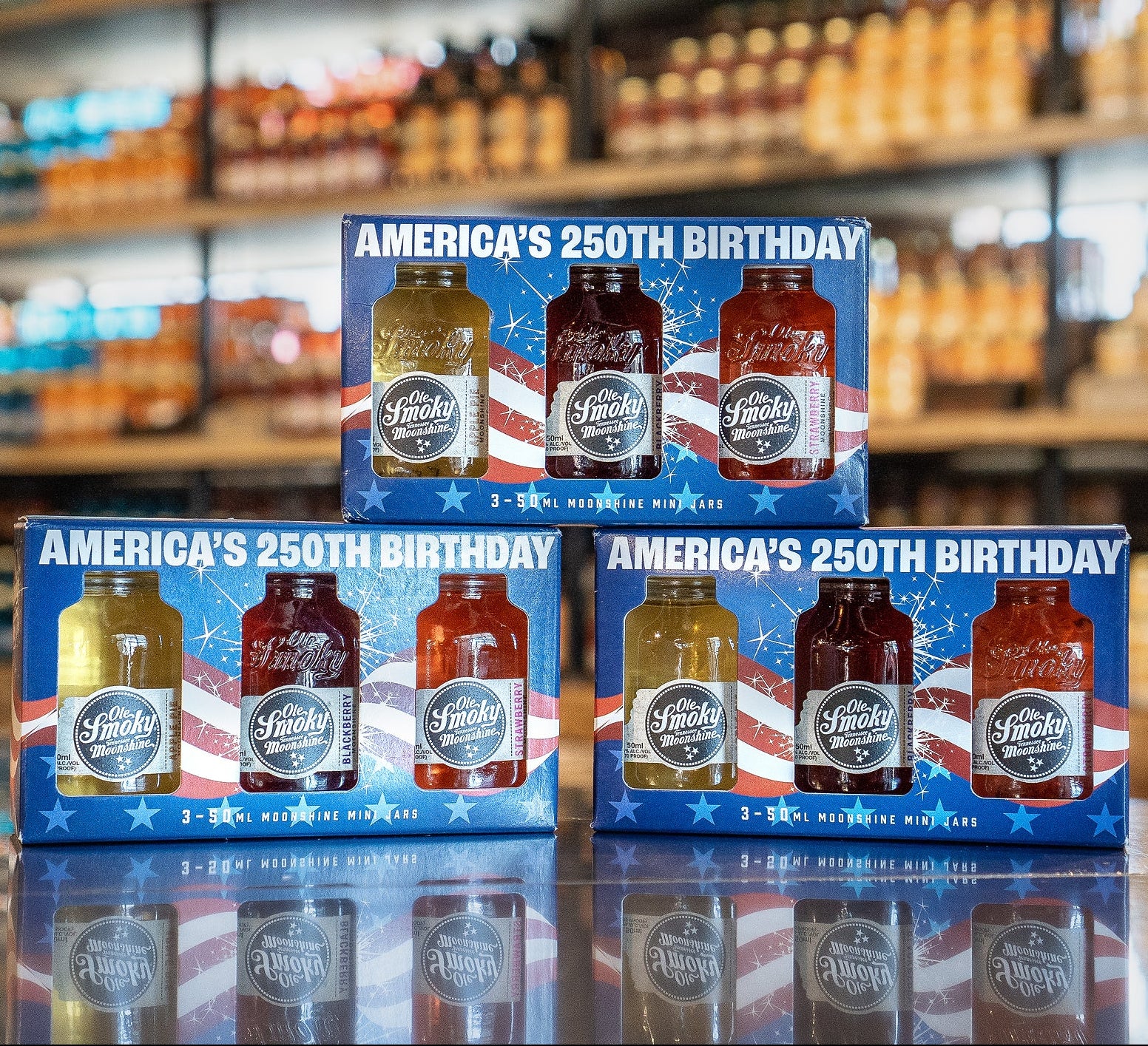 Set of four boxes celebrating America's 250th birthday with bottles on a blurred store shelf background.