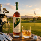 A bottle of Penelope Architects of Golf whiskey stands on a wooden table at a golf course, with a filled golf bag, white golf glove, and a glass of amber whiskey beside a white golf ball. The background shows a golf green and distant hills under a warm sunset sky.