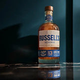A bottle of Russell's Reserve 13 Years Old Kentucky Straight Bourbon Whiskey stands on a reflective dark surface against a deep teal background. The amber liquid inside the clear glass bottle is visible, with the white and blue label clearly showing the brand name.