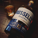 A bottle of Russell's Reserve Kentucky Straight Bourbon Whiskey with a 13-year-old label, shown on a dark wooden surface next to a glass of the amber whiskey.