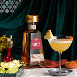 1800 Tequila bottle with a cocktail garnished with a pineapple slice on a dark green curtain background.