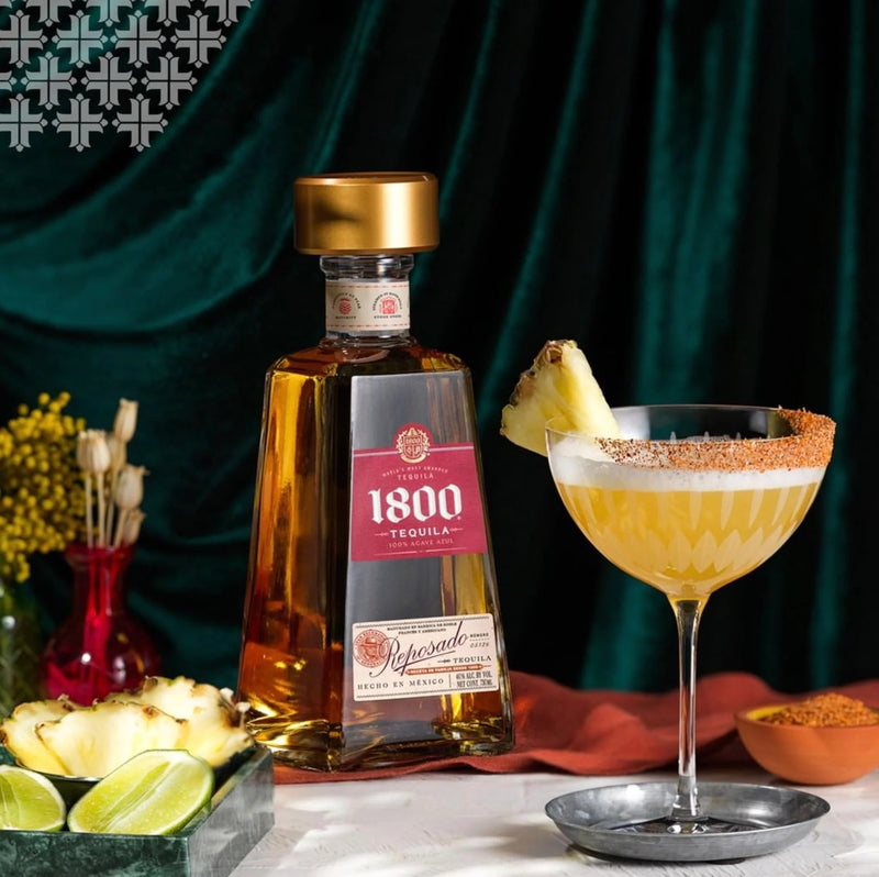 1800 Tequila bottle with a cocktail garnished with a pineapple slice on a dark green curtain background.