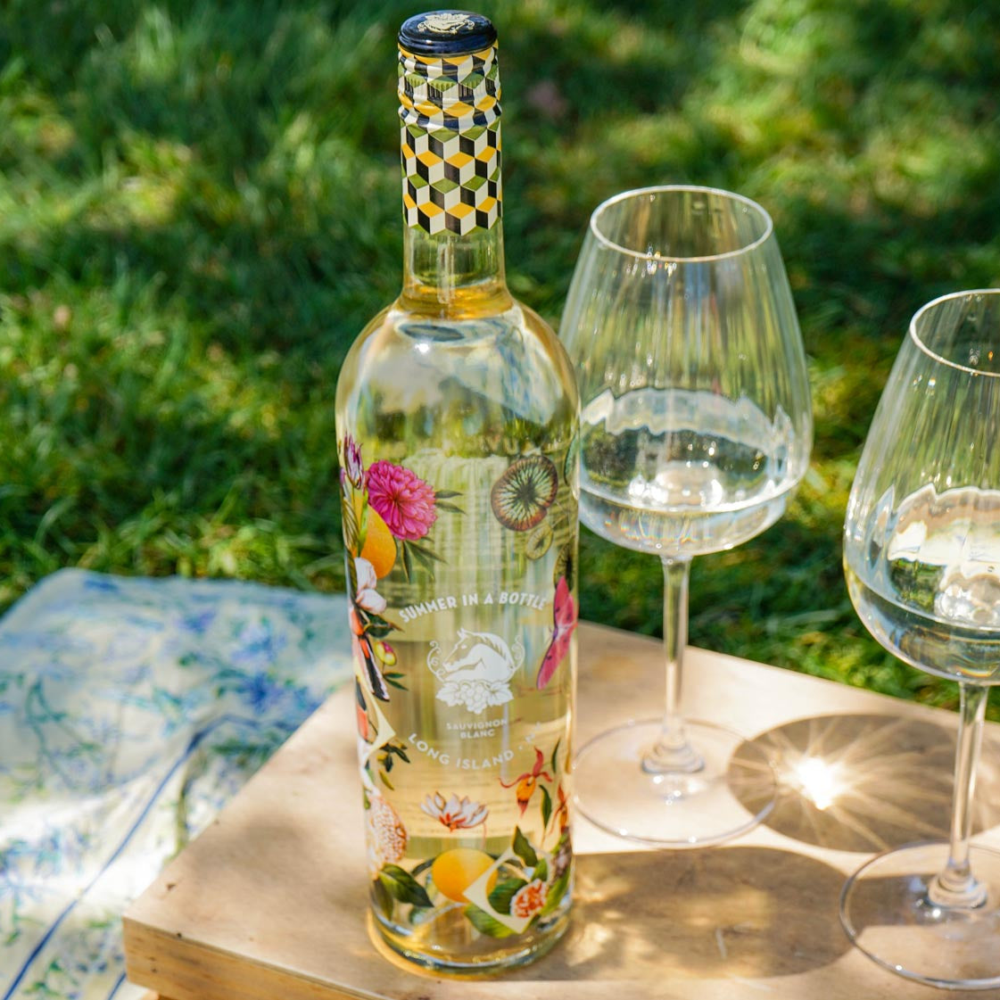 Bottle of floral-patterned wine with two glasses on a wooden table outdoors.