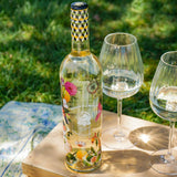 Bottle of floral-patterned wine with two glasses on a wooden table outdoors.