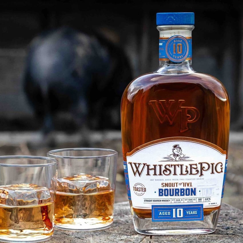 WhistlePig bourbon bottle with two glasses of whiskey on a rustic surface.