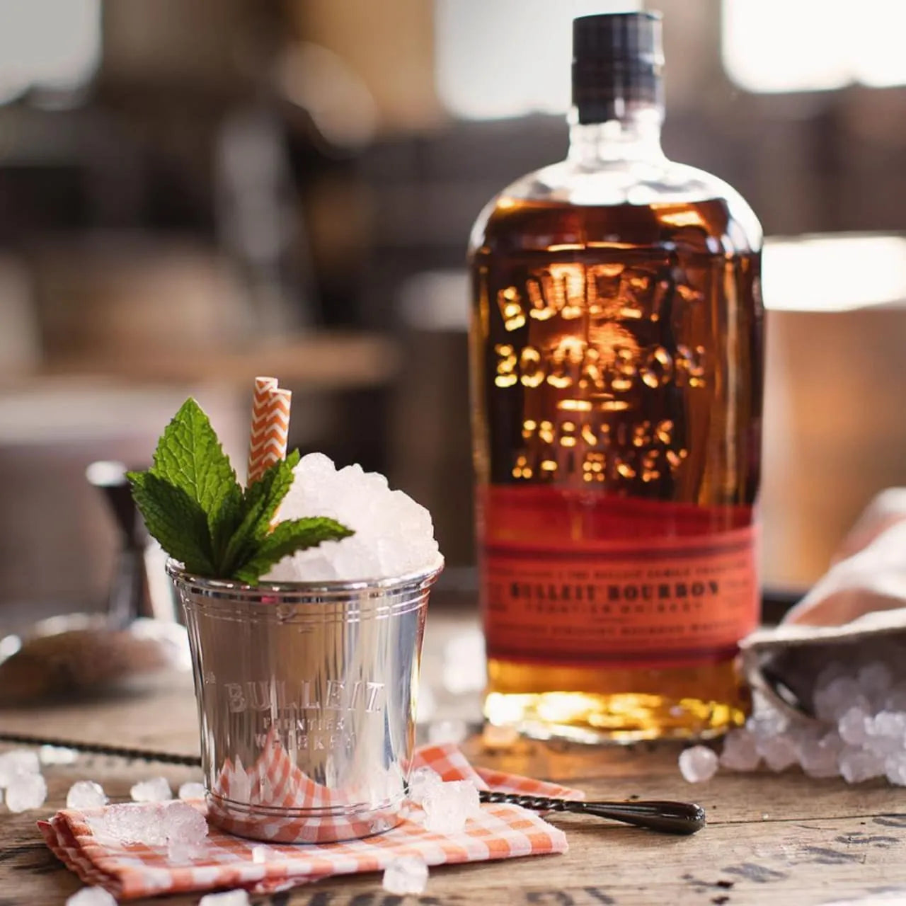 Bulleit Bourbon bottle next to a mint julep cocktail with crushed ice and fresh mint, served in a silver cup on a rustic bar setup.