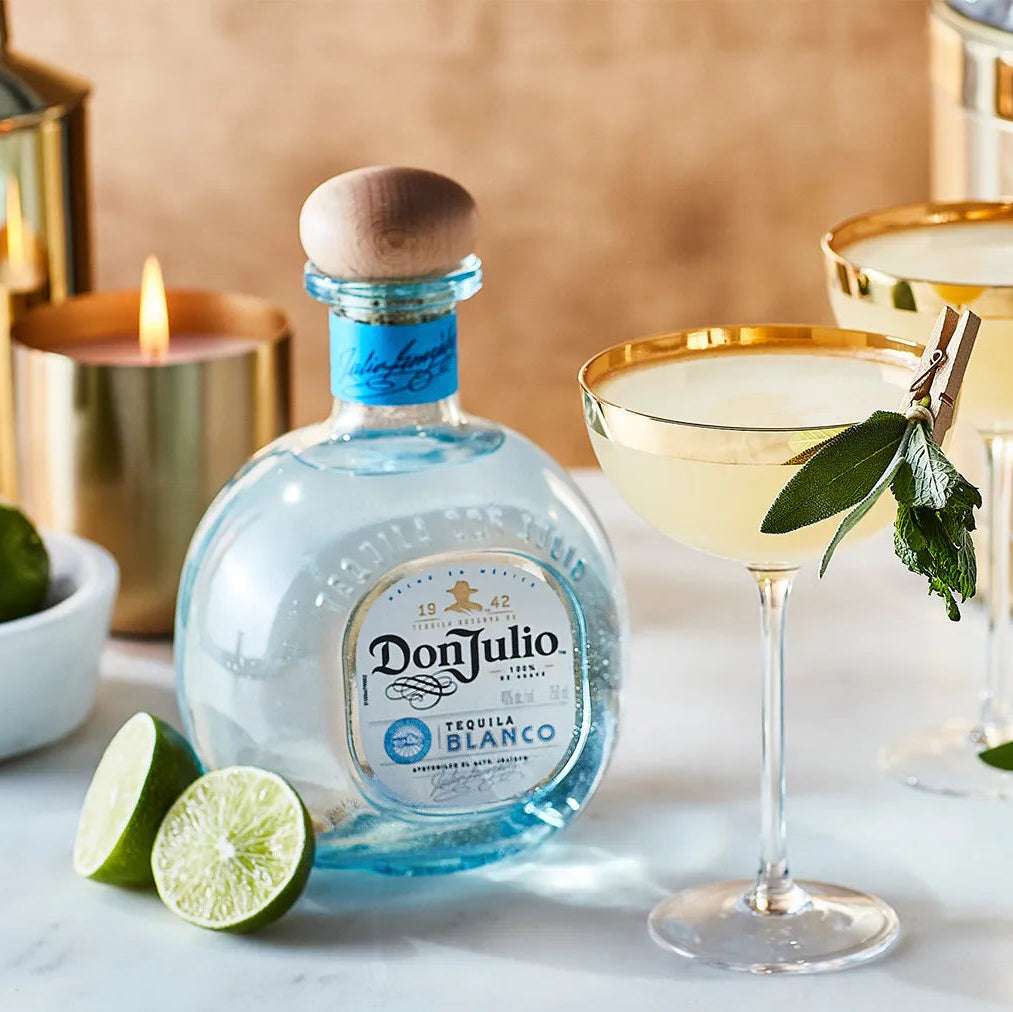 Don Julio Blanco Tequila bottle with two golden-rimmed margarita glasses, fresh limes, and cocktail tools in a luxurious bar setting.