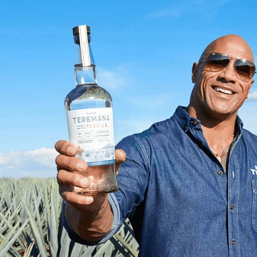 Lifestyle photo of Teremana Blanco Tequila bottle held by The Rock outdoors in an agave field, showcasing small batch handcrafted tequila from Mexico.